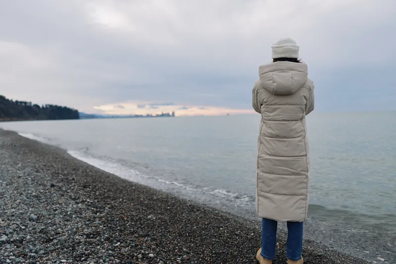 Winter beach coat person sea horizon calm coastline shoreline landscape, a solitary figure stands on a pebbled shore, watching the distant water under a cloudy sky.