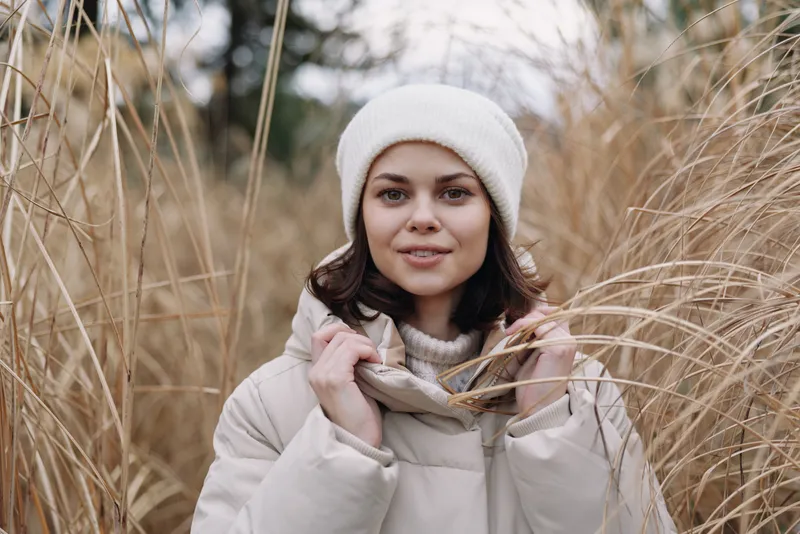 Woman in a light winter coat and knit hat stands among tall dry grass in an open field, calm expression and soft natural light suitable for fashion and lifestyle photography