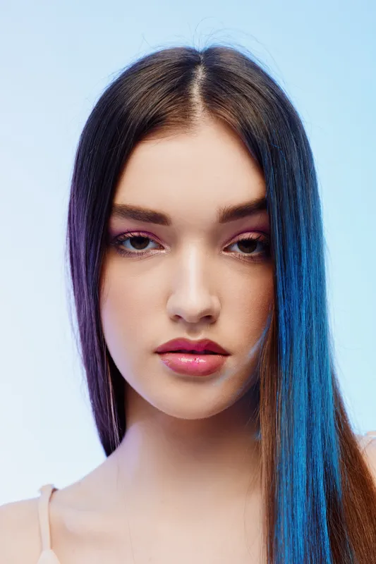 Close-up portrait of a young woman with long straight hair and natural makeup against a light blue background. Concept for beauty, fashion, and skincare.