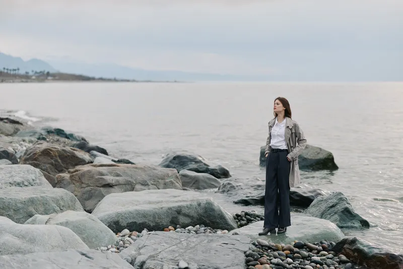 Young woman stands on rocky shore looking at sea wearing casual clothes. Overcast sky and calm water create peaceful mood. Concept for nature, solitude, and contemplation.