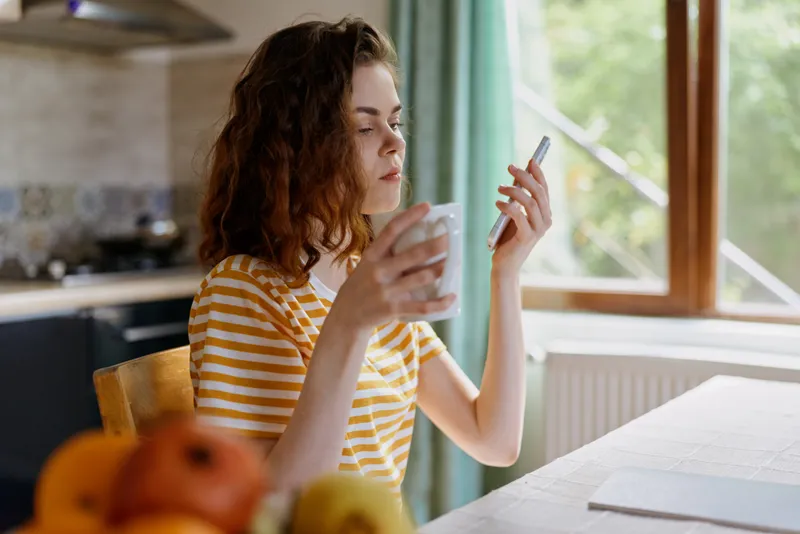 Woman in a striped shirt sits at a bright kitchen table, looking at a smartphone while holding a cup. Casual home moment highlighting daily routine and relaxed wellness. Soft light enhances warmth