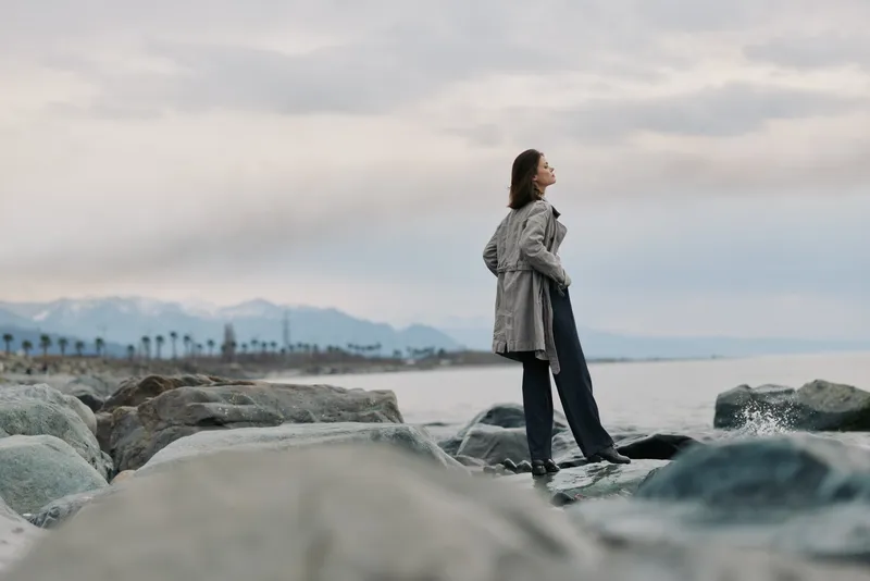 Woman in coat stands on rocky shore looking at cloudy sky with distant mountains and palm trees. Concept of contemplation, nature, and peaceful outdoor moment.