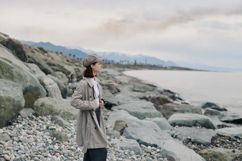 Young woman in grey coat and cap stands on rocky beach looking at sea. Overcast sky and distant mountains create calm, reflective mood. Concept of solitude, nature, and contemplation.
