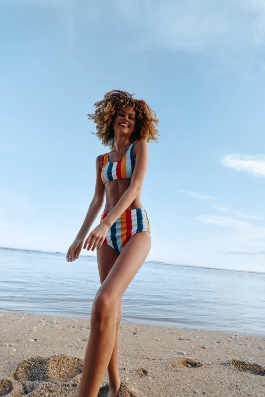 Beach scene shows a young woman in a striped bikini enjoying a sunny day by the ocean, embodying a carefree moment at the shoreline.