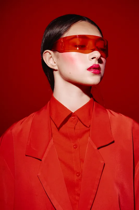 Fashion model in red suit wears oversized tinted sunglasses against red background. Bold makeup and sleek hairstyle create a modern beauty editorial concept.