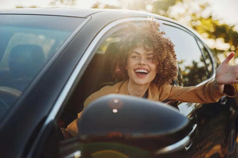 Bright, joyful woman in a car leans out the window, sunlit outdoor scene, casual style, candid lifestyle shot with retro film color and warm glow.