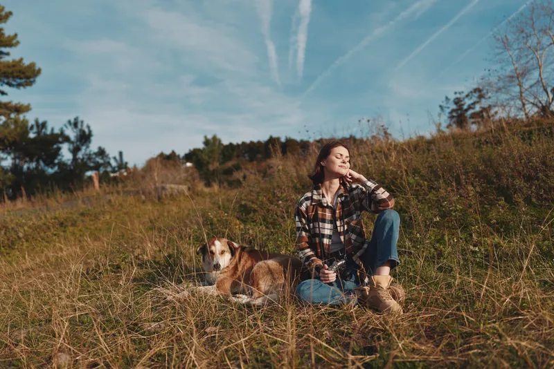 Woman with a dog resting in an open field under a blue sky, enjoying a calm moment in nature, wearing a plaid shirt and jeans during a sunny afternoon.