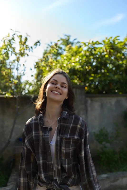 Smiling woman outdoors in a plaid shirt, captured in a sunny, relaxed lifestyle moment amid greenery and blue sky, conveying natural confidence and carefree charm