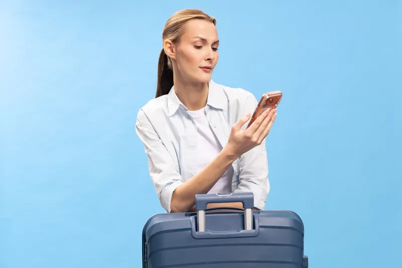 Young woman with suitcase looks at smartphone, preparing for travel. Casual outfit and blue background create concept of vacation, trip planning, and modern lifestyle.