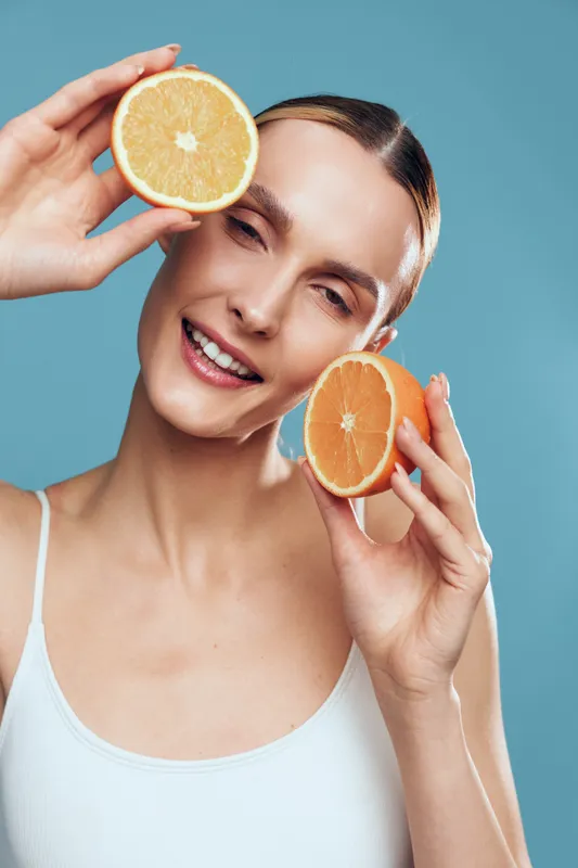 Healthy young woman in white tank top holds orange slices near her eyes, smiling on blue background, vitamin C wellness and skincare concept using fresh fruit for natural brightening of skin