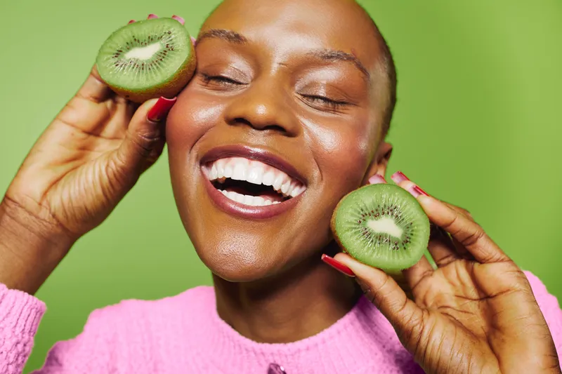 Happy black woman with dark skin holds two halves of fresh kiwi near face, smiling with closed eyes on green background. Concept of healthy lifestyle, fruit, and natural beauty.