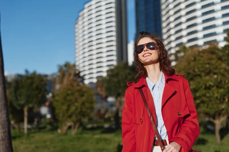 Young woman in a red coat walks outdoors in a sunny park with modern buildings behind, smiling confidently, enjoying a casual urban moment.