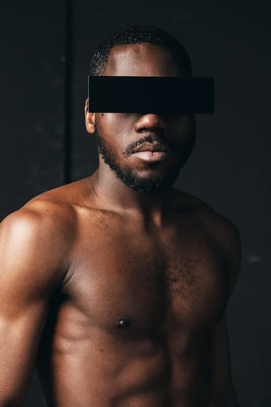 African man with muscular body and dark skin posing shirtless in creative conceptual visual style with black censor bar on eyes for anonymity and artistic expression.