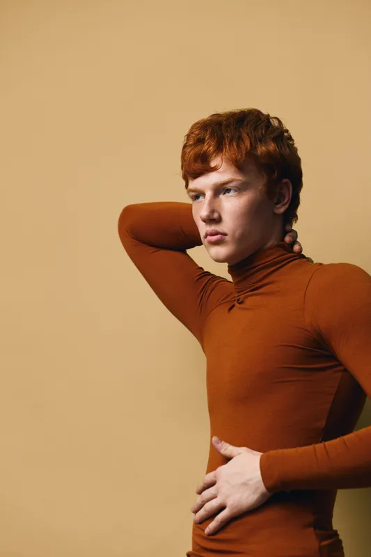 Young man with red hair wearing brown turtleneck posing with hand on neck and waist against beige background. Studio portrait with thoughtful expression and casual style.