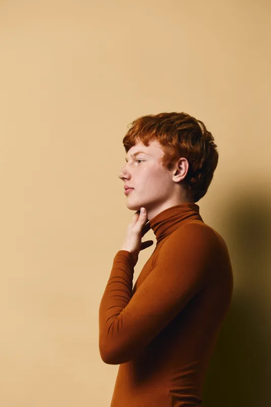 Portrait of a redhead young man wearing brown turtleneck, posing in profile against beige background with short hair and thoughtful expression in studio lighting.