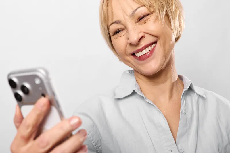 Elderly woman smiling with smartphone in studio white background, taking selfie, making video call, cheerful expression, modern communication technology concept