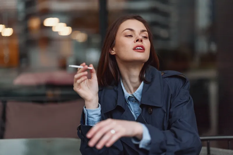 Young woman with brunette hair sits at an outdoor cafe, smoking a cigarette, wearing a denim jacket and blue shirt, a candid urban moment.