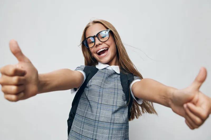 Girl with glasses smiling and showing thumbs up gesture wearing backpack dress on light background. Happy child portrait with long hair expressing positivity and confidence.