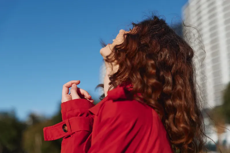 Young woman with curly red hair wearing a red jacket looks up thoughtfully against a clear blue sky in an urban environment.