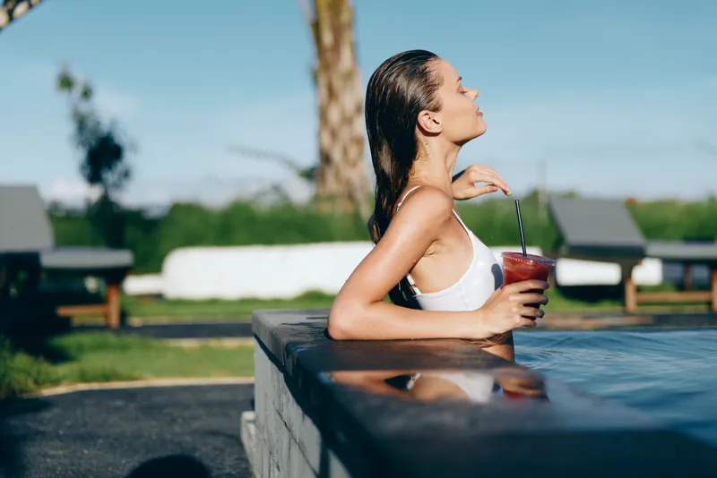 Young woman with wet hair leans on a pool edge, sipping a red beverage through a straw, enjoying a sunny outdoor moment beside a tidy backyard pool.