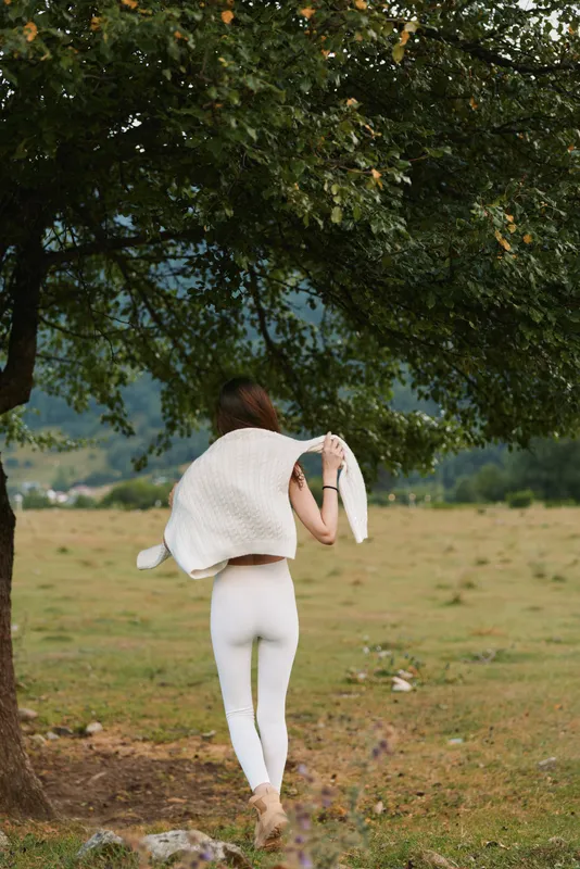 Young woman walking away in white leggings with a light shawl over her shoulders through a sunlit meadow under a leafy tree and distant hills frame the scene softly.