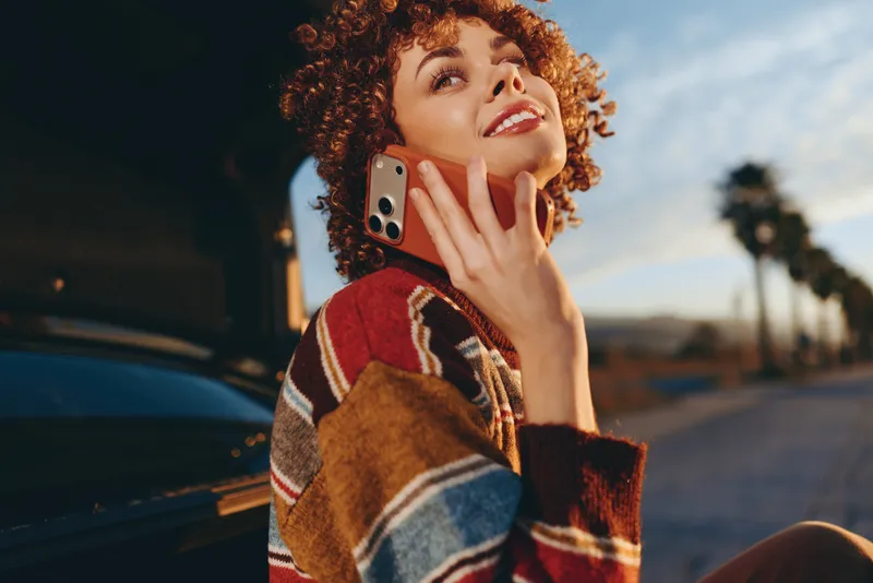 Woman lifestyle outdoors, smiling portrait by car, curly hair, holding phone near cheek, warm sunset light, relaxed posture, casual sweater, urban street background