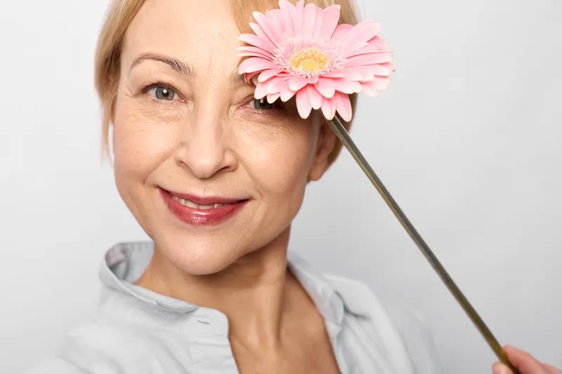 Woman, beauty, skincare, flower, closeup, smile, face, healthy, wellness, spa, mature portrait with pink blossom near eye, suggests self care, hydration and calm