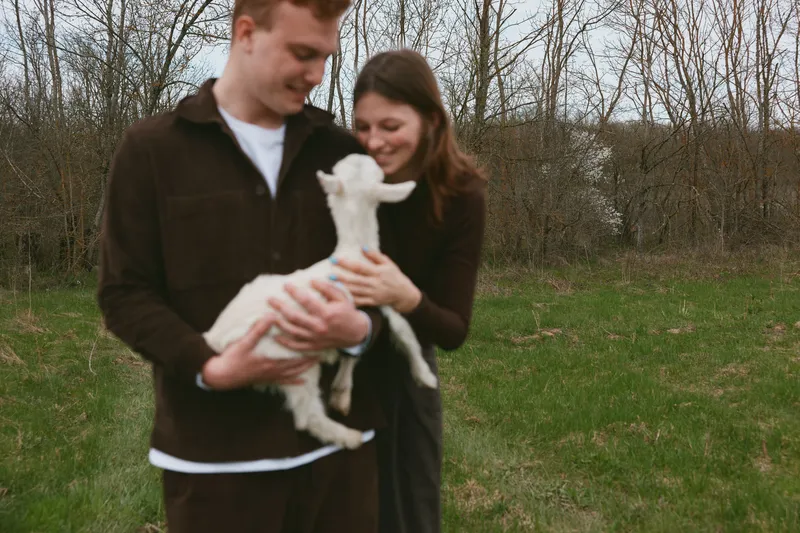 A smiling couple cradles a small white goat kid in a grassy field, enjoying a gentle farm moment outdoors. A warm lifestyle scene focused on animals and rural life.