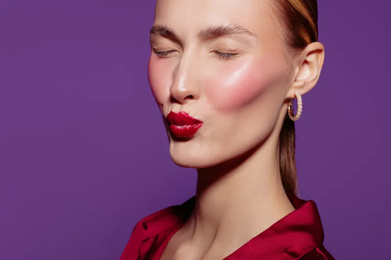 Close-up of a young woman with red lipstick and earrings making a kiss face with closed eyes. Purple background and red blouse create a vibrant and playful mood for beauty concept.