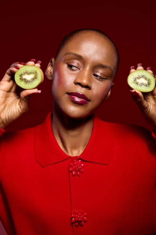 African woman with dark skin holds two kiwi halves near face wearing red shirt on red background. Concept of healthy eating, fresh fruit, natural vitamins, and vibrant lifestyle.