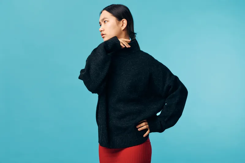 Woman wearing black sweater and red skirt posing with hand on hip looking to side isolated on blue background casual fashion style portrait with thoughtful expression.