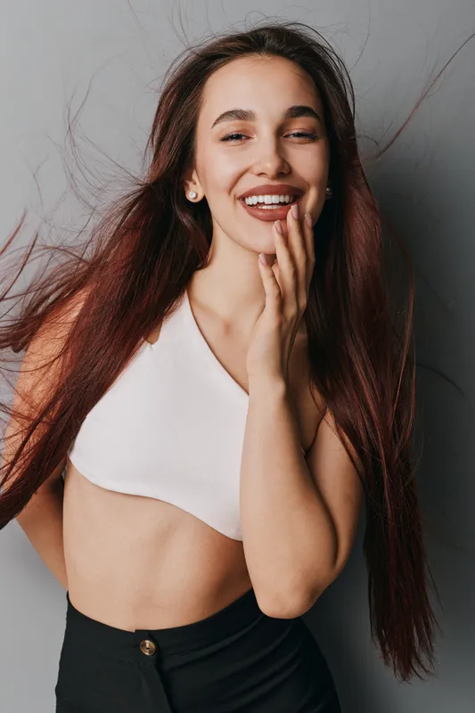 Young woman with long hair smiling and touching face in studio. Casual outfit with white top and black pants, natural makeup, confident and happy expression for lifestyle portrait.