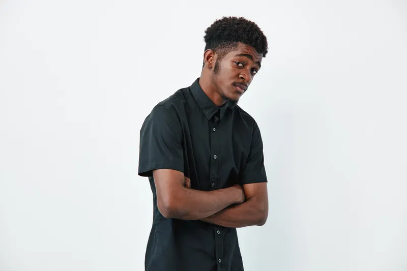 Young African American man with dark skin and curly hair wearing black shirt standing with arms crossed looking sideways on white background. Casual style and confident pose.