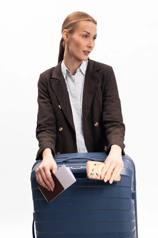 Young woman in brown jacket holds passport and smartphone while standing behind blue suitcase on white background. Travel concept with modern tourist preparing for trip and journey essentials.