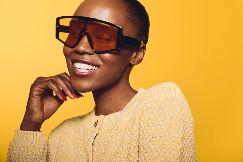 Smiling African woman with dark skin and short hair wears large brown sunglasses and a textured yellow sweater. Studio portrait on yellow background with bright, cheerful mood and fashion concept.