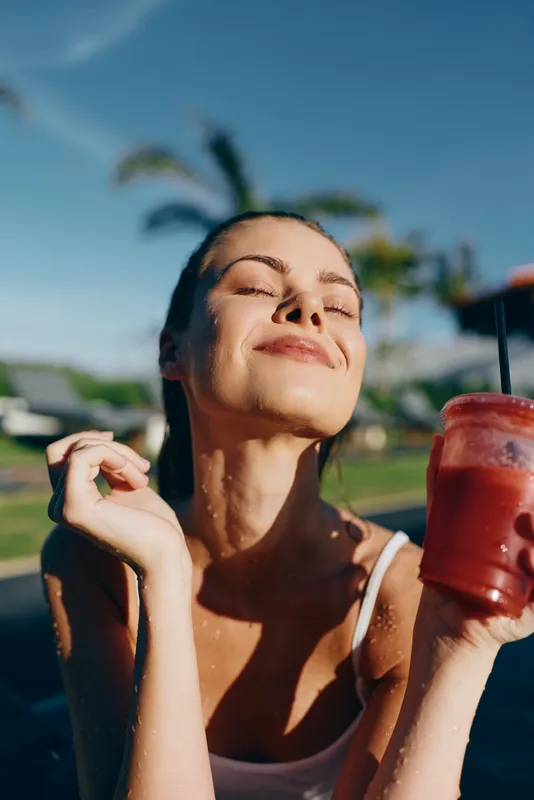 A woman outdoors in summer sunlight enjoys a refreshing smoothie beverage by the poolside, eyes closed, smile radiating relaxation and happiness in a carefree lifestyle scene.