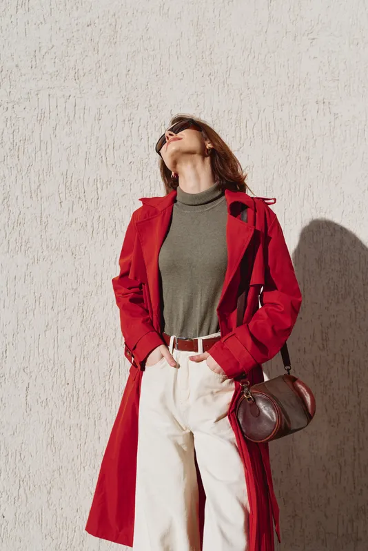 A stylish woman in a red coat poses against a textured wall, head tilted back and hands tucked in her pockets, showcasing a bold fall look and confident mood.