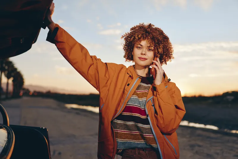 woman, lifestyle, phone, curly, hair, jacket, car, sunset, outdoors, young woman in orange jacket stands beside car holding phone to ear, curly hair blowing, warm evening sky, travel lifestyle moment