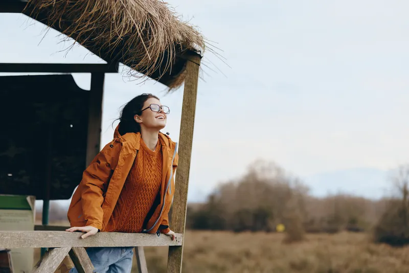 Woman with glasses, wearing jacket, standing outdoors under rural umbrella of wooden straw, smiling while leaning on wooden railing, spring countryside background, lifestyle portrait