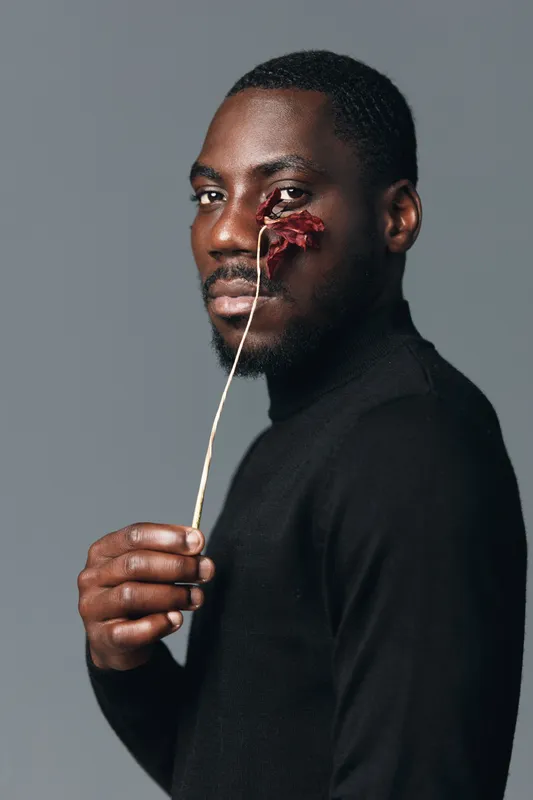 African man with dark skin holding dried flower near face in creative conceptual visual style. Portrait of black male model with thoughtful expression and artistic mood on gray background.