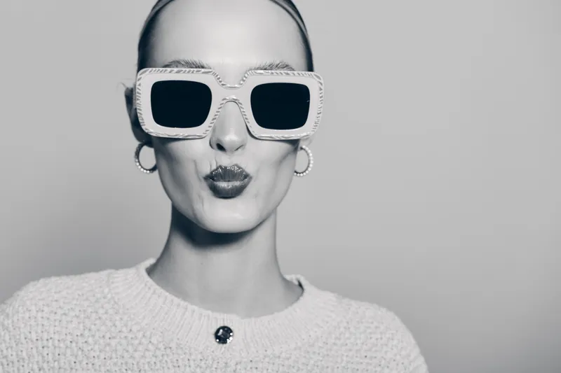 Young caucasian woman with pale skin wearing large sunglasses and knitted sweater makes a kiss face. Minimal studio background with fashion and beauty concept in black and white.