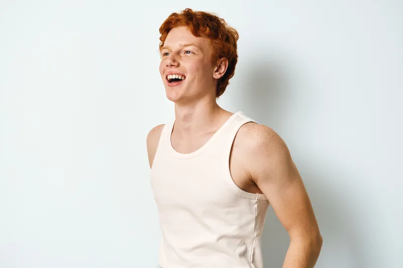 Young man with red hair laughing and wearing sleeveless shirt standing against white background. Casual, happy and confident smiling male in bright studio with natural expression.