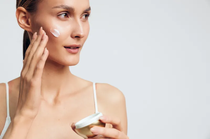 Young woman applies facial cream on cheek holding jar with skincare product isolated on white background with soft natural light and calm expression.