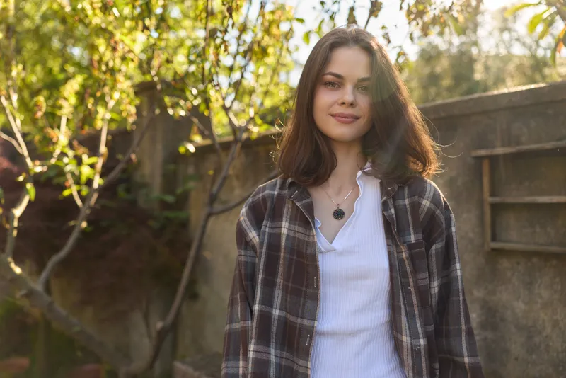 Young woman with brown hair stands in a sunlit garden wearing a plaid shirt over a white blouse, smiling softly as warm light creates a relaxed outdoor mood