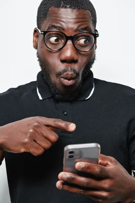 African man with dark skin and glasses looking surprised while pointing at smartphone screen. Casual black shirt, facial hair, and expressive face on white background.
