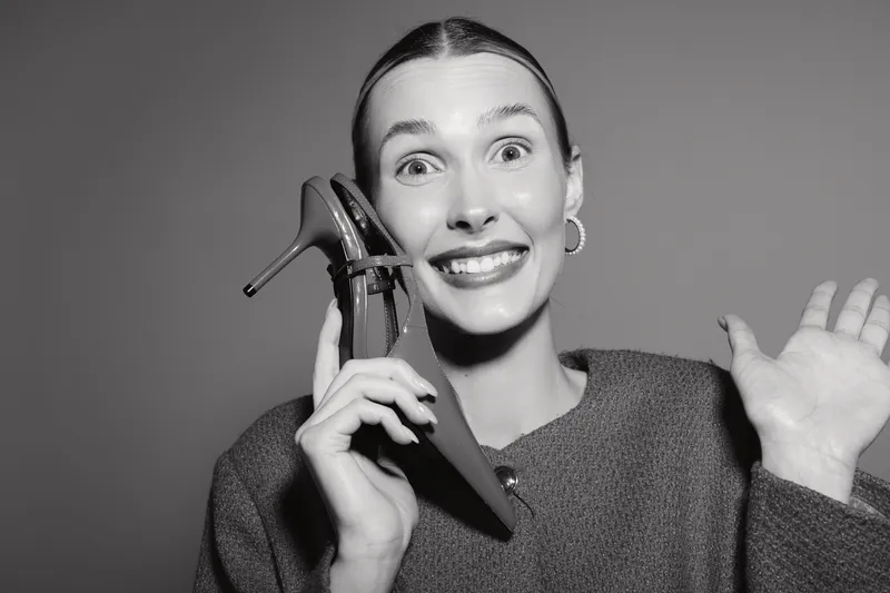 Smiling white woman holds red shoe near face and waves hand on grey background. Concept for fashion and happiness.