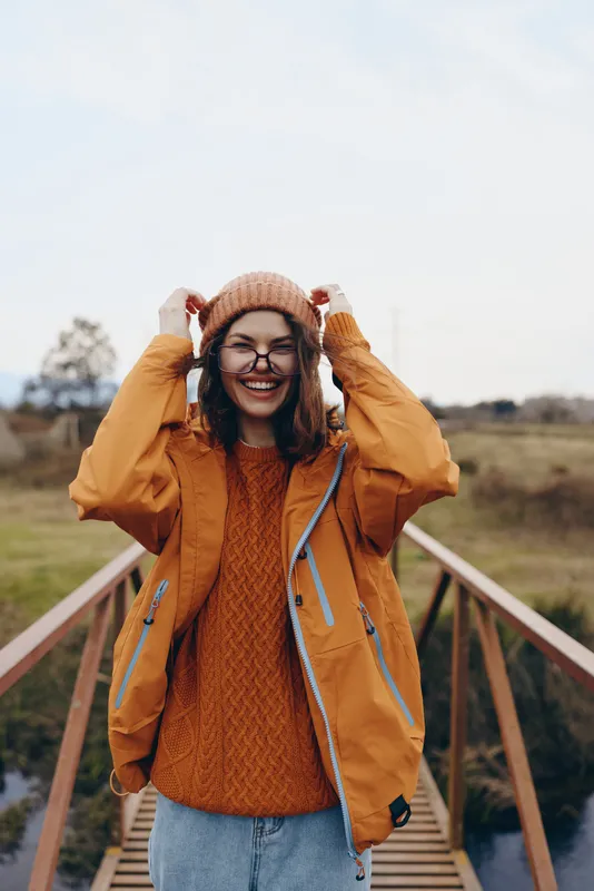 Woman outdoors lifestyle, smiling while adjusting a knit hat and orange jacket on a wooden bridge. Countryside background, soft daylight. Travel autumn mood, happiness today.