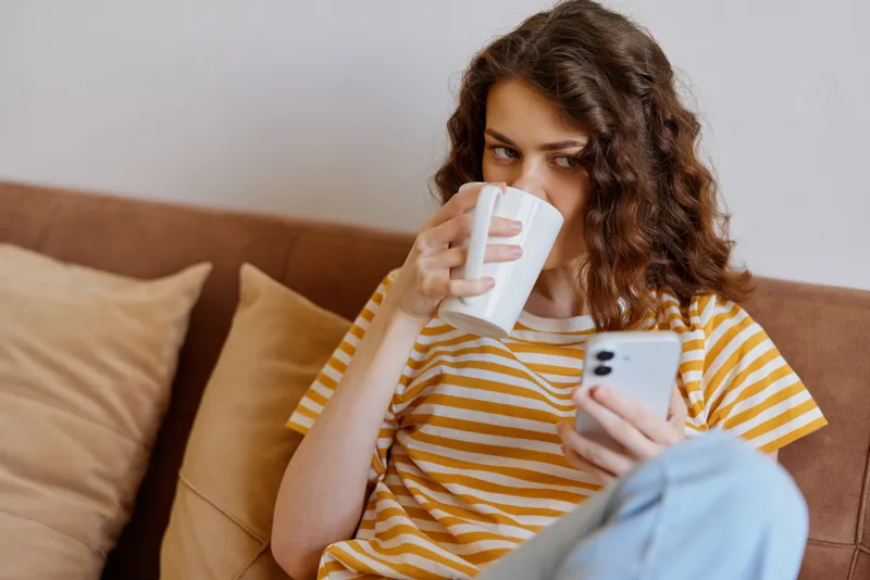 Young woman sits on a cozy sofa at home, sipping from a mug while browsing on her smartphone, creating a relaxed intimate moment with soft lighting and casual style.
