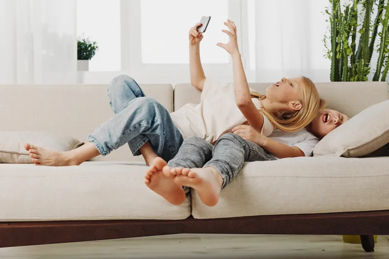 Two children lying on a beige sofa playing with a smartphone, laughing and having fun in a bright living room. Concept of family joy, sibling bonding, and leisure time at home.