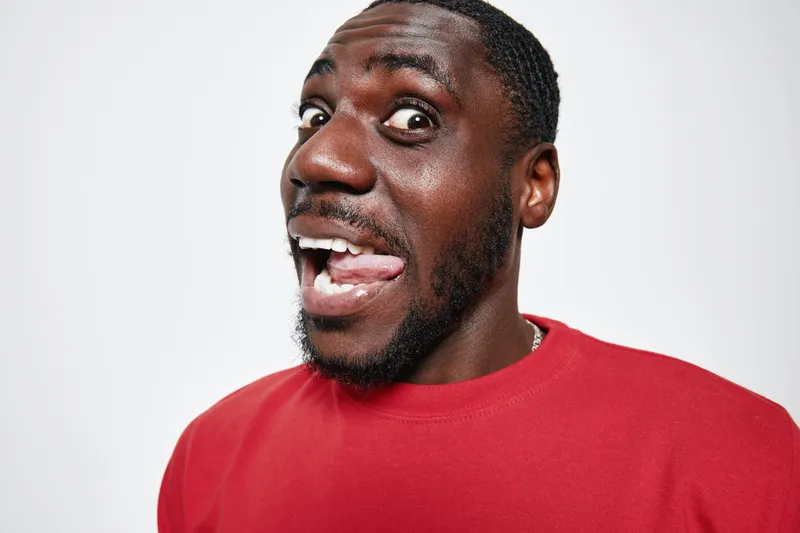 African American man with dark skin wearing red sweater making funny face with tongue out and wide eyes on white background in studio portrait.